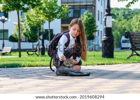 Cute little girl in school uniform and with backpack stroking a stray cat on the street, in the first time goes to school. 