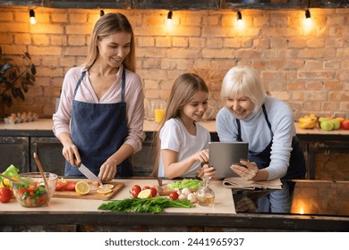 Cute little girl showing some to her mother and grandmother in the tablet while preparing fresh vegetables in kitchen with light bulb on the background. Looking recipe to the internet - Powered by Shutterstock