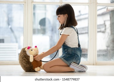 Cute little girl sit on warm home floor play doctor patient game listen with stethoscope  to fluffy hedgehog heart, small kid future nurse involved in childish activity take care of stuffed toy