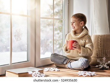 Cute little girl sitting by the window, drinking warm beverage and looking at the winter forest. Child makes paper snowflakes for decoration windows. Kid enjoys the snowfall.