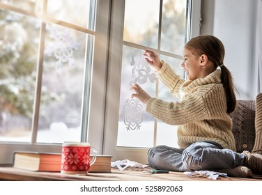Cute little girl sitting by the window and looking at the winter forest. Child makes paper snowflakes for decoration windows. Kid enjoys the snowfall.