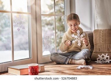 Cute Little Girl Sitting By The Window And Looking At The Winter Forest. Child Makes Paper Snowflakes For Decoration Windows. Kid Enjoys The Snowfall.