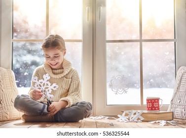 Cute Little Girl Sitting By The Window And Looking At The Winter Forest. Child Makes Paper Snowflakes For Decoration Windows. Kid Enjoys The Snowfall.