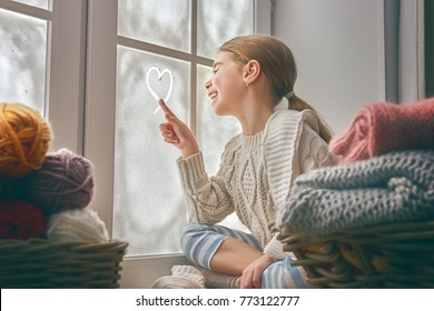 Cute Little Girl Sitting By The Window And Drawing Heart On Frozen Glass. Kid Enjoys The Winter.