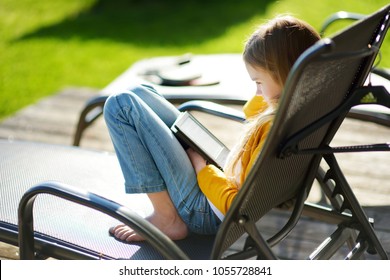 Cute Little Girl Sitting In A Garden And Reading An Ebook On Sunny Summer Evening. Education For Kids.