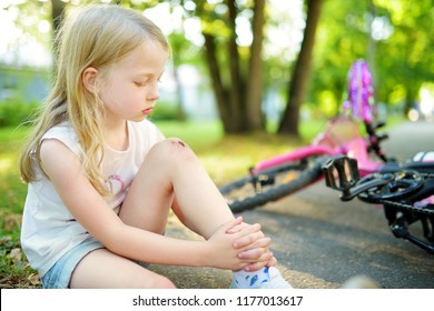 Cute Little Girl Sitting On The Ground After Falling Off Her Bike At Summer Park. Child Getting Hurt While Riding A Bicycle. Active Family Leisure With Kids.