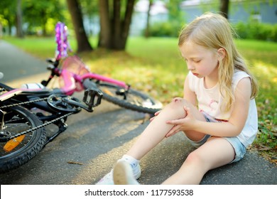 Cute Little Girl Sitting On The Ground After Falling Off Her Bike At Summer Park. Child Getting Hurt While Riding A Bicycle. Active Family Leisure With Kids.