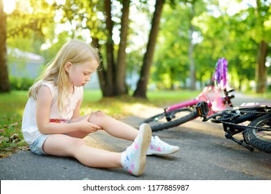 Cute Little Girl Sitting On The Ground After Falling Off Her Bike At Summer Park. Child Getting Hurt While Riding A Bicycle. Active Family Leisure With Kids.