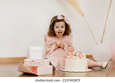 A cute little girl is sitting on the floor with a cake and gifts on her birthday and eating a lollipop
