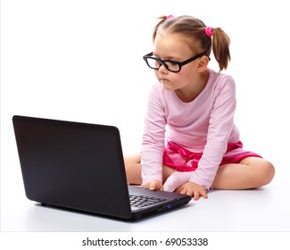 Cute little girl is sitting on floor with her laptop, wearing glasses, isolated over white