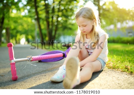 Cute little girl sitting on the ground after falling off her scooter at summer park. Child getting hurt while riding a kick scooter. Active family leisure with kids.