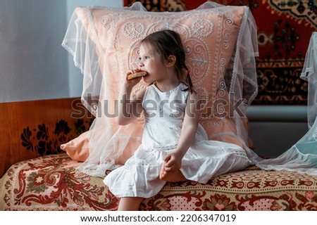 A cute little girl is sitting on the bed and eating bread. She is sitting on the bed in the atmosphere of the former USSR