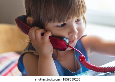 A Cute Little Girl Is Sitting At A Table And Talking On An Old Red Home Landline Phone. Communication, The Concept Of Relationships. European Children, Baby Calls On The Phone.