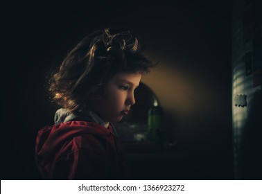 A Cute Little Girl Is Standing In A Dark Room And Looking Away. Reflection Of Light, Back Light, Shadow. Fear And Dark. Beautiful Indigo Child. Low Key Photography.