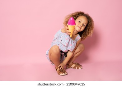 Cute little girl in summer clothing with ice cream sitting on the floor isolated over pink background.