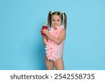 Cute little girl in swimsuit with drink on light blue background