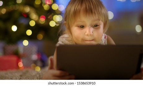 Cute Little Girl With Tablet Computer In Her Hands Lies On The Carpet Under The Christmas Tree.