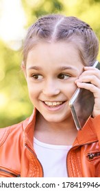 Cute Little Girl Talking. Hold Phone Near Face. Young Woman Smile. Not Looking At Camera. Outdoor School. Green Background. Smiling Speach. Happy Expression. Student Lifestyle. Vertical