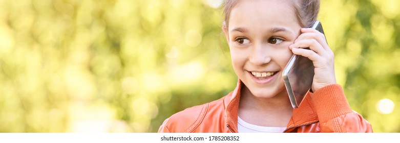 Cute Little Girl Talking. Hold Phone Near Face. Young Woman Smile. Not Looking At Camera. Outdoor School. Green Background. Smiling Speach. Happy Expression. Student Lifestyle. Horizontal Banner