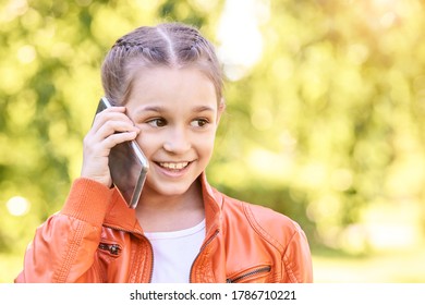 Cute Little Girl Talking. Hold Phone Near Face. Young Woman Smile. Not Looking At Camera. Outdoor School. Green Background. Smiling Speach. Happy Expression. Student Lifestyle