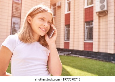 Cute Little Girl Talking. Hold Phone Near Face. Young Woman Smile. Not Looking At Camera. Outdoor School. Green Background. Smiling Speach. Happy Expression. Student Lifestyle