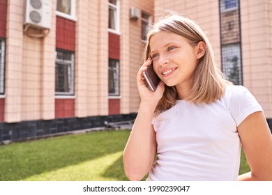 Cute Little Girl Talking. Hold Phone Near Face. Young Woman Smile. Not Looking At Camera. Outdoor School. Green Background. Smiling Speach. Happy Expression. Student Lifestyle