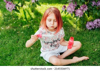 Cute Little Girl With Toned Red Hair Blowing Soap Bubbles While Sitting On A Green Lawn In The Garden. Having Fun During School Holidays At Home. Offline Activity For Kids. Happy Childhood Concept.