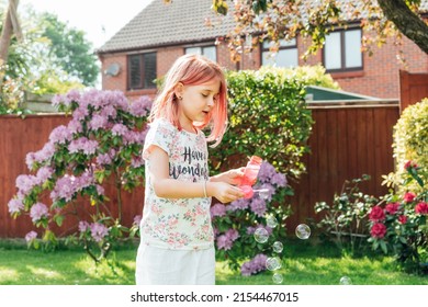 Cute Little Girl With Toned Red Hair Blowing Soap Bubbles While Playing On A Green Lawn In The Garden. Having Fun During School Holidays At Home. Offline Activity For Kids. Happy Childhood Concept.