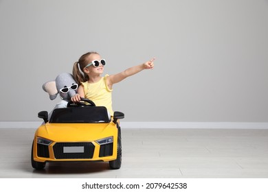 Cute little girl with toy elephant driving children's car near grey wall indoors. Space for text