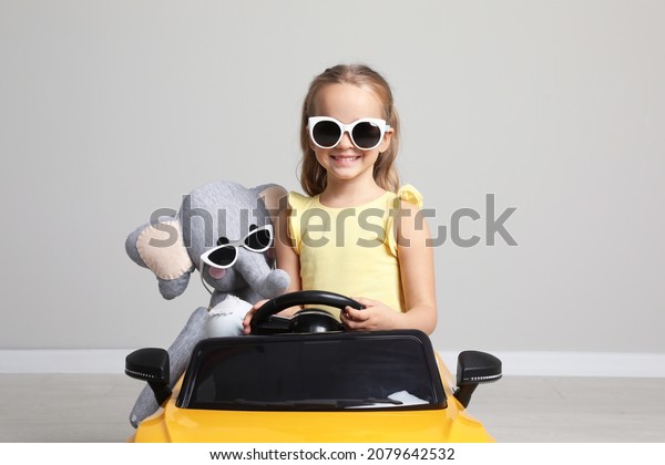 Cute little girl with toy elephant driving\
children\'s car near grey wall\
indoors