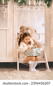 A cute little girl unwraps a Christmas gift while sitting in the children's room on a decorated bed. New Year's Holiday - Shutterstock ID 2396436207