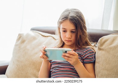 Cute little girl using technology device sitting on sofa. Girl playing on phone.