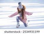 Cute little girl in warm pink coat, grey knitted hat and scarf, snood learning to skate on ice skating rink in park. Fall down and have fun. Winter sport, outdoors. Holiday and seasonal concept.