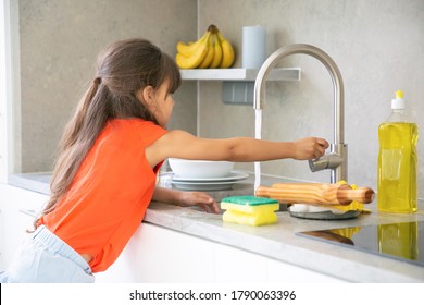 Cute Little Girl Washing Dish In Kitchen By Herself. Child Reaching Kitchen Sink Faucet Tap And Turning On Water. Domestic Shores Concept