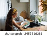 Cute little girl washing dishes with sponge in kitchen sink at home. Child doing chores concept.