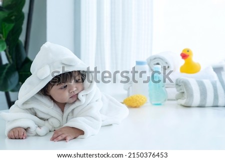 Cute little girl wearing a bathrobe relaxes on the table after taking a bath, yellow duck toy and bath accessories in blurred background, happy little girl at home