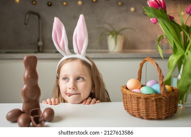 Cute Little Girl Wearing Bunny Ears Eating Chocolate Easter Rabbit. Kid Playing Egg Hunt On Easter. Adorable Child Celebrate Easter At Home.