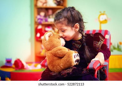 cute little girl in wheelchair telling her secret to plush bear in kindergarten for kids with special needs