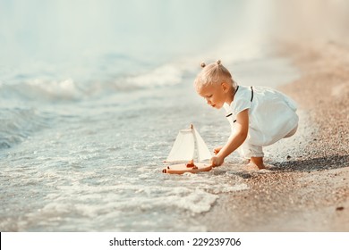 A cute little girl in white clothes playing with a toy ship on the beach on a warm sunny summer day. Holidays at sea. Funny kids