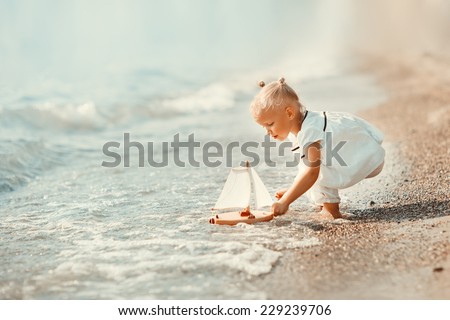 A cute little girl in white clothes playing with a toy ship on the beach on a warm sunny summer day. Holidays at sea. Funny kids