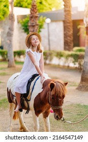 a cute little girl in a white dress and straw hat sits on top of a pony. equestrian sport for children.