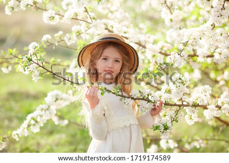 a cute little girl in a white dress and a straw hat smiles in a blooming garden in spring