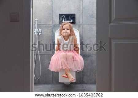 Cute little girl in white pink dress sitting on toilet on background of walls with gray tiles, view from open door.