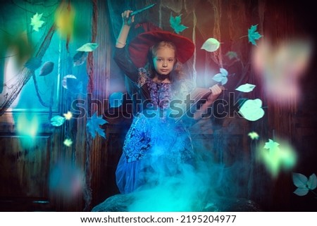 Cute little girl in a witch costume conjures over a burning cauldron with a book of spells and magic wand in her hands. Halloween time. Old house interior with leaf fall around. 