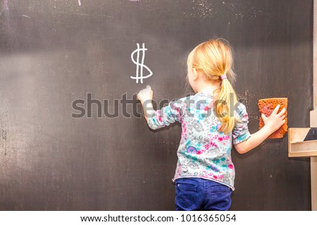 Cute little girl writing Dollar Sign on chalkboard in a classroom