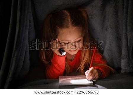 Cute Little Girl Writing In Notebook While Hiding Under Blanket With Flashlight, Smiling Happy Preteen Female Child Holding Torch In Darkness, Filling Diary Or Drawing In Notepad, Closeup