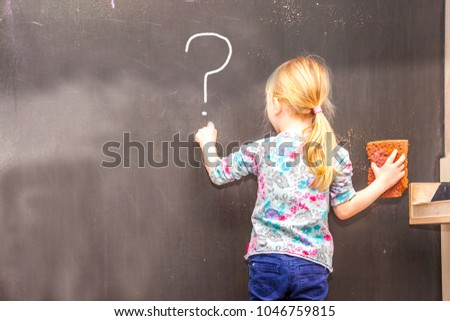 Cute little girl writing question mark on chalkboard in a classroom