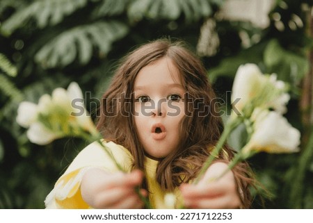 Cute little girl in a yellow dress, holding spring flowers in her hands, standing against the background of leaves,