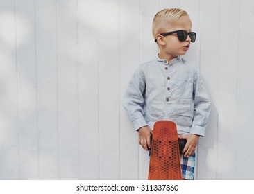 Cute little guy in big trendy sunglasses that he has borrowed from his mother or father posing with his skateboard against a white wooden wall with copyspace