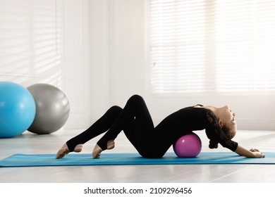 Cute little gymnast with ball stretching on mat indoors